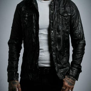 Może przedstawiać: Czarna skórzana koszula zapinana na guziki z dwiema kieszeniami na piersi. Koszula jest noszona na białym t-shircie i czarnych spodniach. Skóra ma teksturowany, pomarszczony wygląd. Koszula ma klasyczny kołnierz i mankiety zapinane na guziki.