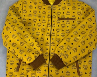 May include: Yellow bomber jacket with a repeating black MCM logo pattern. The jacket features a brown collar, cuffs, and hem, with a full-length zipper. A small pocket is visible on the chest.