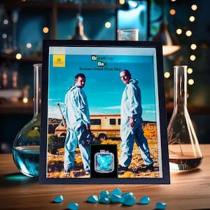 Peut inclure: Une œuvre encadrée représentant la série Breaking Bad. L'image montre deux hommes en combinaisons bleues, avec le texte "Breaking Bad Screen Used Blue Sky". Un petit récipient de bonbons bleus est également visible, ainsi que plusieurs morceaux éparpillés sur la table.