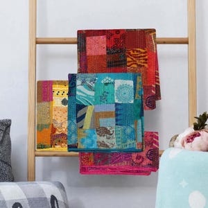 May include: Three rectangular fabric quilts featuring patchwork designs with visible running stitch patterns. One quilt displays red and orange tones, another features blue and teal shades, and the third showcases yellow and orange patterns.