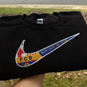 May include: Black crewneck sweatshirt featuring a large embroidered Nike swoosh design. The swoosh incorporates the FCB logo and the phrase "més que un club" in blue and yellow. The sweatshirt is made of a soft material.