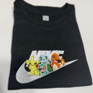 May include: Black t-shirt with a white embroidered Nike logo featuring a group of colorful cartoon characters. The characters include a yellow Pikachu, a blue Squirtle, a green Bulbasaur, and an orange Charmander.