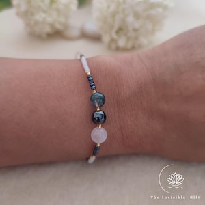 Op de afbeelding: Een delicate kralenarmband met een reeks kleine witte, donkergrijze en goudkleurige kralen, gecentreerd met een grotere mosagaat kraal, een donkere metalen kraal en een lichtroze rozenkwarts kraal, met de tekst The Invisible Gift.