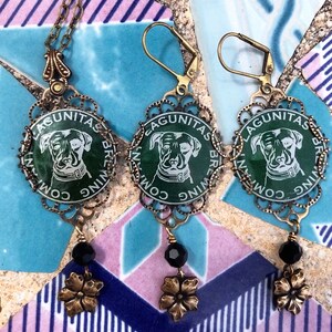 May include: A pair of earrings featuring a green and white Lagunitas Brewing Company bottle cap with a dog illustration. The earrings have black beads and gold flower accents.