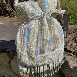 Può includere: Una borsa a sacchetto bianca con coulisse, ricoperta da intricate perline, paillettes iridescenti, perle e lunghe frange di perline sul fondo. La borsa presenta un manico intrecciato e un'etichetta scura con il testo Folklore Kingdoms.