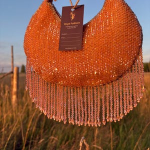 Op de afbeelding: Een halvemaanvormige handtas bedekt met glinsterende oranje kralen met een lange kralenfranje langs de onderrand. Een donkerbruin rechthoekig label is bevestigd, met de tekst Royal Folklore, Inspired by Kingdoms, crafted for you, Product, Price en Handmade in India.