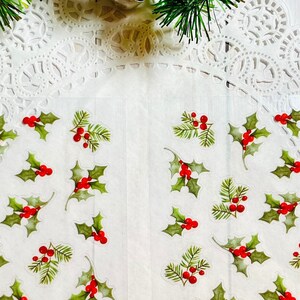 May include: Transparent stickers with green holly leaves and red berries, displayed on a white doily. The stickers are spread across the surface, with a sprig of evergreen and a red and white string in the background. A festive, holiday theme.
