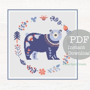 May include: A cross-stitch pattern featuring a blue bear with floral details, surrounded by a wreath of flowers and leaves. The pattern is titled "PDF Instant Download" and is from "See Spot Create".