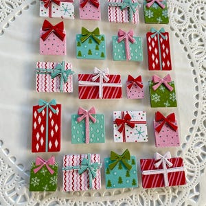 May include: A collection of colorful, miniature gift boxes with various patterns and bows. The boxes feature designs like snowflakes, Christmas trees, and zigzags. Bow colors include red, pink, and green. The gifts are arranged on a white, lacy doily.