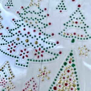 May include: A sheet of clear adhesive-backed paper with various Christmas tree and snowflake designs. The designs are made of small, glittery, red, green, and gold dots.