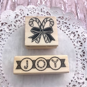 May include: Two wooden Christmas stamps. The first stamp features a pair of candy canes tied with a bow. The second stamp features the word "JOY" with each letter in a circle with a candy cane design.
