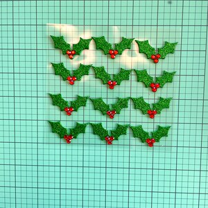 May include: A selection of glittery green holly leaf embellishments with red berry accents. These festive decorations are arranged on a grid, ideal for holiday crafts. Each holly leaf is approximately 2.5 cm wide.