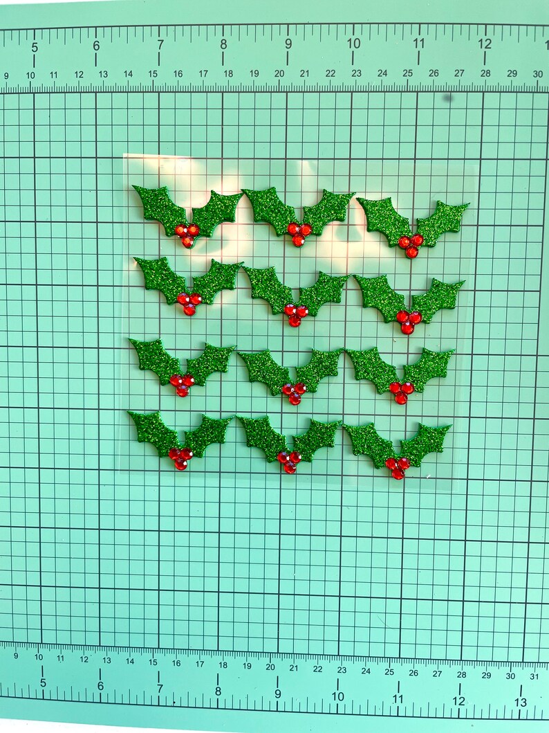 May include: A selection of glittery green holly leaf embellishments with red berry accents. These festive decorations are arranged on a grid, ideal for holiday crafts. Each holly leaf is approximately 2.5 cm wide.