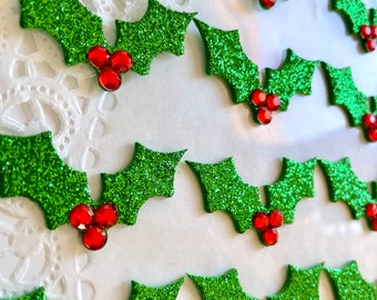 May include: Close-up of several glittery green holly leaf decorations with red jewel berries. The leaves are arranged in rows, creating a festive and decorative pattern. Perfect for holiday crafts and embellishments.