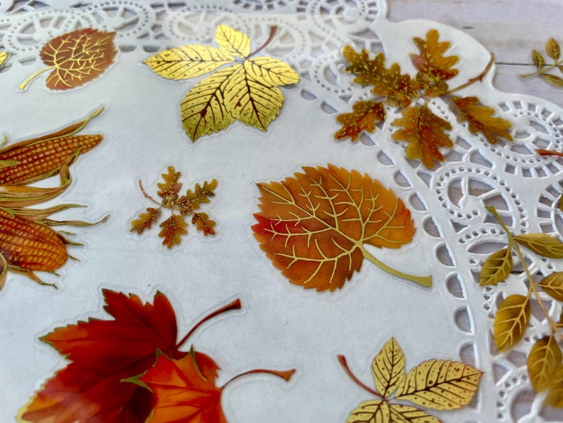 Pu&ograve; includere: Una collezione di adesivi a tema autunnale con varie forme di foglie nei toni dell'oro, del rosso e dell'arancione. Alcuni adesivi hanno dettagli in lamina d'oro. Gli adesivi sono disposti su una superficie bianca bordata di pizzo.