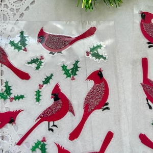 May include: A sheet of red cardinal stickers with holly leaves. The cardinals are sparkly and have black eyes and beaks. The holly leaves are green with red berries.