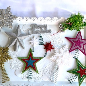 May include: Assortment of sparkly Christmas tree toppers in various shapes and colors, including stars, snowflakes, and a Christmas tree. The toppers are displayed on a white tray with a lace doily.