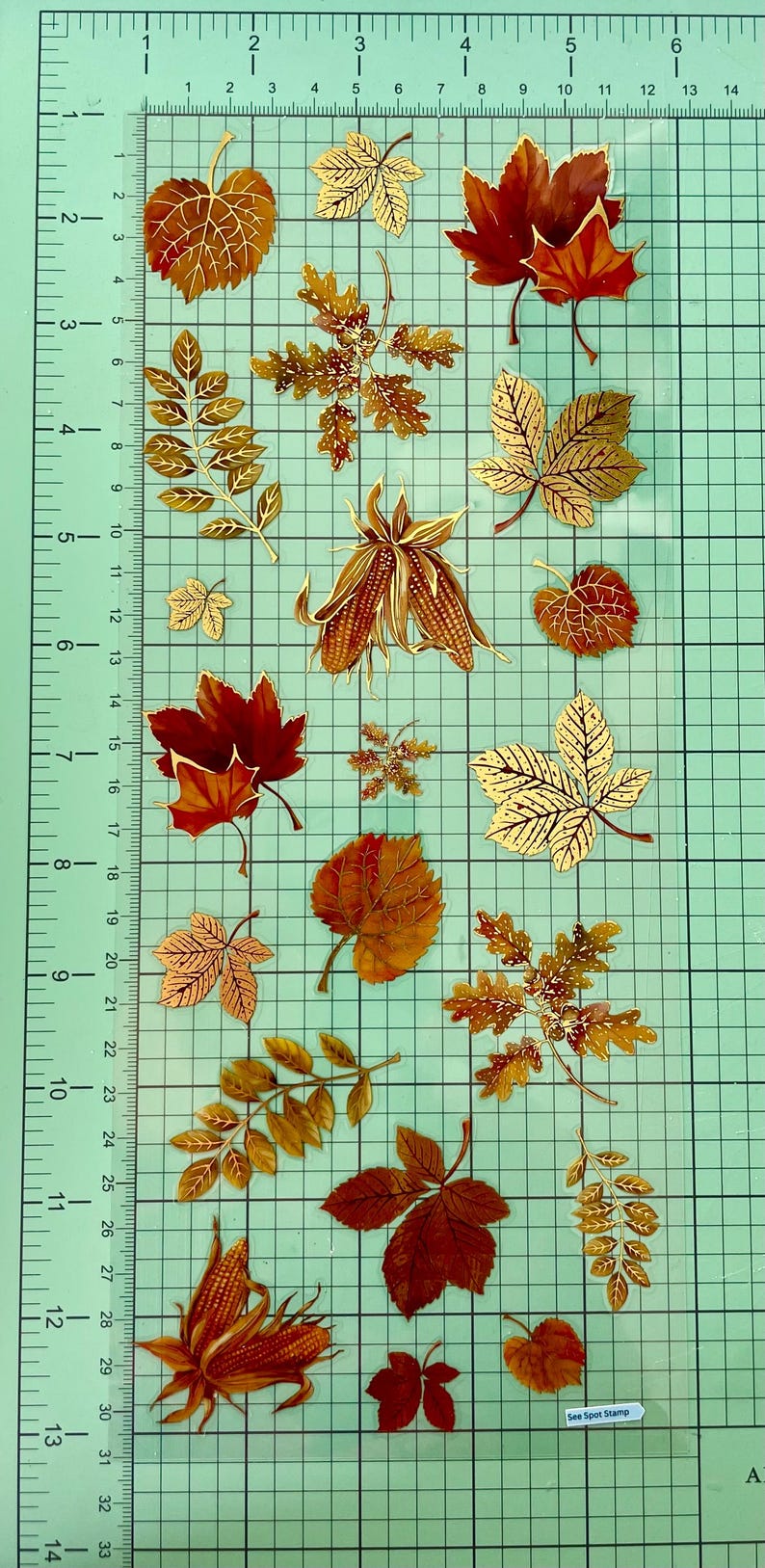 Pu&ograve; includere: Una collezione di adesivi a tema autunnale con vari disegni di foglie nei toni dell'oro, del rosso e del marrone. Gli adesivi includono foglie d'acero, foglie di quercia e pannocchie di mais. Gli adesivi sono su uno sfondo a griglia.