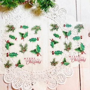 May include: Transparent stickers with holly leaves, red berries, and the words "Merry Christmas" in red script. The stickers are displayed on a white doily, with a sprig of evergreen in the background.