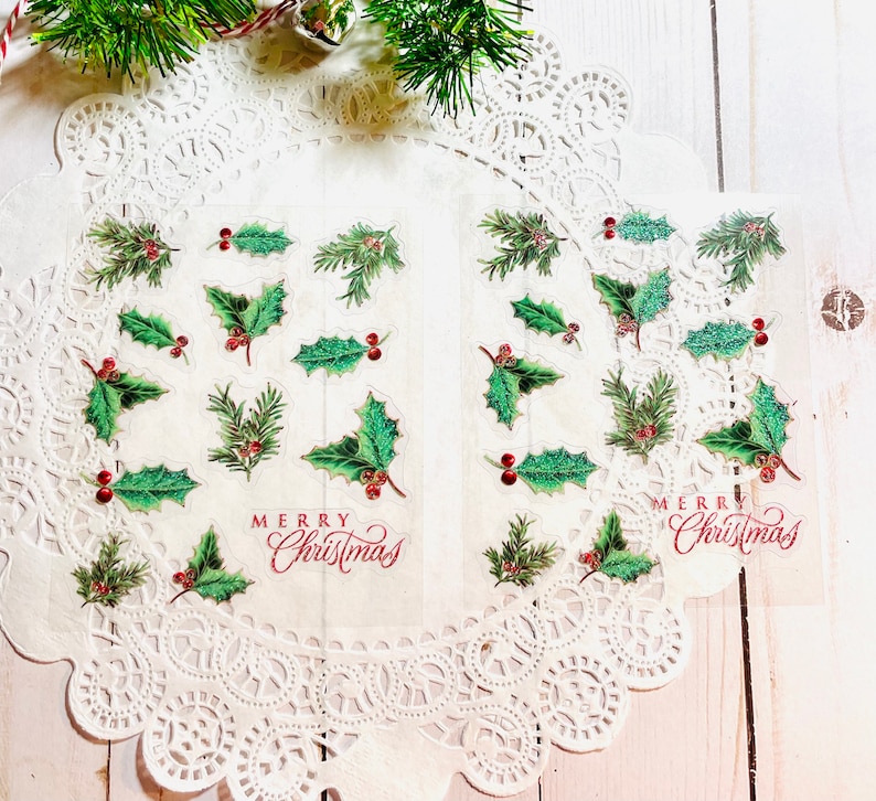 May include: Transparent stickers with holly leaves, red berries, and the words "Merry Christmas" in red script. The stickers are displayed on a white doily, with a sprig of evergreen in the background.