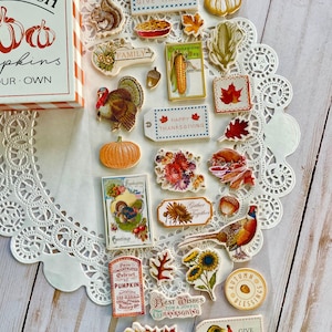 May include: A collection of Thanksgiving themed paper craft embellishments, including a turkey, pumpkins, corn, sunflowers, and leaves. The embellishments are on a white doily and a wooden surface. The text on the embellishments includes "Give Thanks", "Happy Thanksgiving", "Gather Together", and "Best Wishes for a Joyful Thanksgiving".