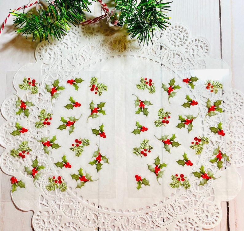 May include: Transparent stickers with green holly leaves and red berries, displayed on a white doily. The stickers are spread across the surface, with a sprig of evergreen and a red and white string in the background. A festive, holiday theme.
