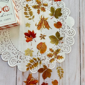 Pu&ograve; includere: Una collezione di adesivi a tema autunnale con vari disegni di foglie nei toni dell'arancione, del rosso e dell'oro. Gli adesivi sono disposti su una tovaglietta bianca con bordi in pizzo, con una piccola scatola che dice "FRESH PUMPKINS FOR OWN".