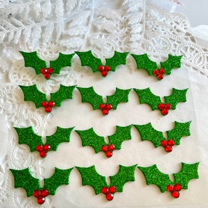 May include: Glittering green holly leaf embellishments with red jewel berries. These festive decorations are ideal for holiday crafts. Each holly leaf measures approximately 3.8 cm wide, adding sparkle to any project.
