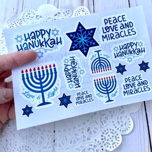May include: A set of blue and white Hanukkah stickers featuring menorahs, stars of David, snowflakes, and the phrase "Happy Hanukkah". Some stickers also include the phrases "Peace Love and Miracles" and "Love & Light".