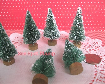 May include: Six miniature artificial Christmas trees with white flocked snow on a white lace doily. The trees are green and have brown bases. The text "See Spot Stamp" is visible in the image.