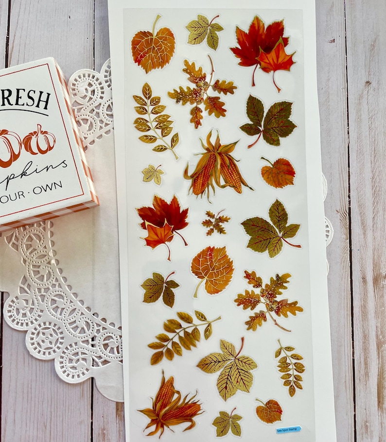 Pu&ograve; includere: Un foglio di adesivi a tema autunnale con vari disegni di foglie nei toni del rosso, dell'arancione e del verde. Gli adesivi raffigurano foglie d'acero, foglie di quercia e altri fogliami autunnali. &Egrave; visibile una piccola scatola con la scritta "FRESH PUMPKINS".