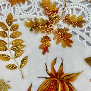 Pu&ograve; includere: Primo piano di adesivi a tema autunnale con illustrazioni dettagliate di foglie e mais. Le foglie sono in tonalit&agrave; arancioni, marroni e dorate, con intricati dettagli delle venature. Gli adesivi sono disposti su uno sfondo bianco e merlettato.