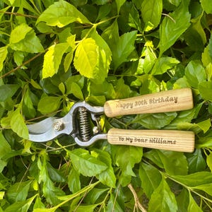 Puede incluir: Tijeras de podar con hojas plateadas y mangos de madera, grabadas con "Happy 30th Birthday" y "To My dear Williams". Las tijeras se encuentran sobre un lecho de hojas verdes.