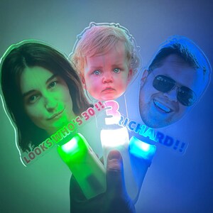 May include: Three light-up face wands with custom photos and text. One wand says "LOOKS WHO'S 30!!", another has the number "3" and the name "RICHARD!!". Each wand has a different coloured light: green, white, and blue.