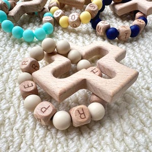 Könnte beinhalten: Holz-Beißring in Sternform mit einem Ring aus cremefarbenen Perlen und Buchstabenwürfeln aus Holz. Andere Beißringe im Bild haben blaue, gelbe und marinefarbene Perlen. Die Beißringe liegen auf einer weißen, strukturierten Oberfläche.
