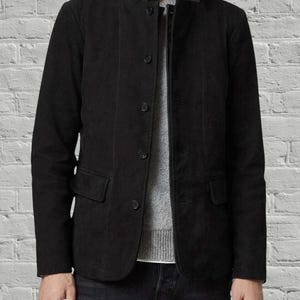Puede incluir: Una chaqueta negra con acabado texturizado, que presenta un cuello alzado con ribete de cuero negro liso, cierre frontal con cinco botones visibles y dos bolsillos con solapa en la parte inferior, sobre un su&eacute;ter de punto gris.