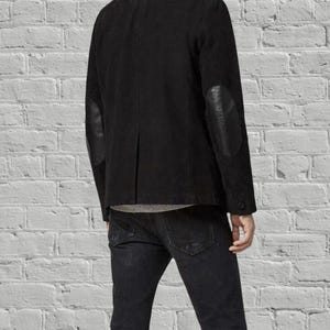 Puede incluir: Una chaqueta de ante negro con cuello alto y parches de cuero liso en los codos de las mangas. La chaqueta se lleva sobre una camiseta gris y se combina con vaqueros de mezclilla gris oscuro, mostrando un corte estructurado y acabado negro.