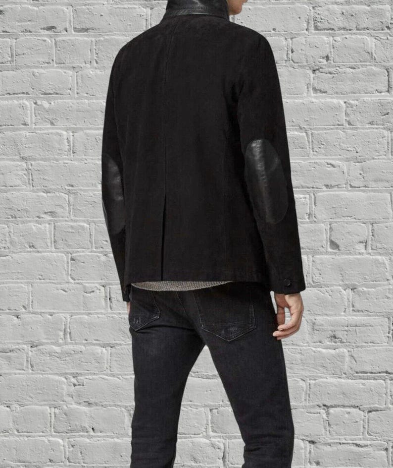Puede incluir: Una chaqueta de ante negro con cuello alto y parches de cuero liso en los codos de las mangas. La chaqueta se lleva sobre una camiseta gris y se combina con vaqueros de mezclilla gris oscuro, mostrando un corte estructurado y acabado negro.