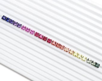 May include: A collection of square-cut gemstones arranged in a gradient of colors. The stones transition from deep blues and purples to pinks, reds, oranges, yellows, and finally, greens. The gems are displayed on a white, striped surface.
