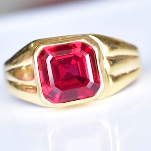 May include: A gold ring with a large, rectangular, faceted red gemstone. The ring has a classic design with a thick band and a setting that holds the gemstone securely. The gemstone reflects light, showcasing its vibrant color and clarity.
