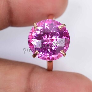 May include: A rose gold ring with a large, round, faceted pink gemstone. The gemstone is secured by four prongs. The ring is set against a blurred background.
