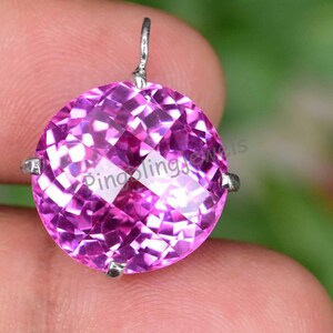 Pode incluir: Um pingente redondo de pedra preciosa rosa facetada com uma argola de prata. O pingente tem um corte brilhante, refletindo a luz. O texto "Pinoolings Jewels" é visível. O fundo está desfocado.