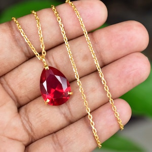 May include: A gold necklace featuring a teardrop-shaped, faceted, red gemstone pendant. The chain is delicate and gold-coloured. The pendant is approximately 3.8 cm long.