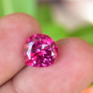 May include: A faceted, heart-shaped, hot pink gemstone. The gem is held in a hand, with a blurred green background. The gem has many facets that reflect light, creating a sparkling effect. The gem is a vibrant shade of pink.