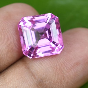 May include: A faceted, rectangular pink gemstone with a brilliant cut. The gem is held in a hand, with a green background. The gem's facets reflect light, creating a sparkling effect. The gem is a vibrant shade of pink.