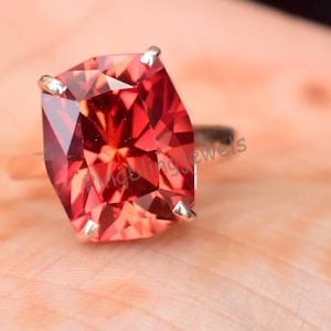 May include: A striking red gemstone ring with a silver band. The cushion-cut gemstone reflects light, creating a dazzling effect. The ring is displayed on a light surface, highlighting the jewellery's beauty.