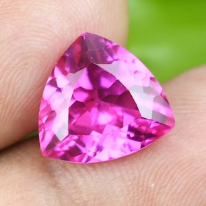 May include: A faceted, triangular-shaped gemstone in a vibrant shade of pink. The gem's multiple facets reflect light, creating a sparkling effect. The gemstone is held in a person's hand, with a green background.
