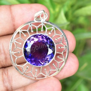 May include: A circular silver pendant with a large, faceted, deep purple gemstone at its centre. The gemstone is set within an openwork silver frame. The pendant has a loop for a chain.