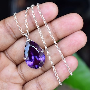 May include: A silver necklace with a large, faceted, teardrop-shaped amethyst pendant. The deep purple gemstone is set in a silver bezel and hangs from a delicate silver chain. The necklace is displayed against a neutral background.