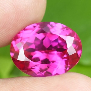 May include: A faceted, oval-cut gemstone in a vibrant magenta hue. The gem's multiple facets reflect light, creating a sparkling effect. The gemstone is held between two fingers against a blurred green background, highlighting its color and clarity.
